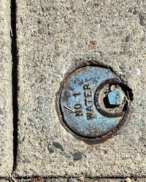 A tan sidewalk with very small inclusions. It is divided into two squares. In the bottom square is a small circle of blue metal that says "No. 1 water". It has a pentagonal drive socket to allow opening it.