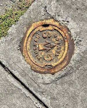 A closeup of a sidewalk oriented diagonally. There are three partial diamonds of dingy cement. A line of short grass runs between two of them. In the bottom diamond is an old round yellow circle of metal with faded yellow paint. Across the middle it says GAS with a raised arrow pointing downward. Around that word it says Bingham Taylor.