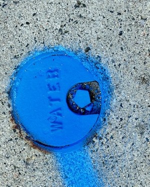 Cemet with many small dark inclusions. Close to the middle is a metal circle painted blue with WATER in a sans serif font across the middle. Below the words is a pentagonal bolt head. A faint blue line of paint goes from the circle across the sidewalk all the way out of the image.