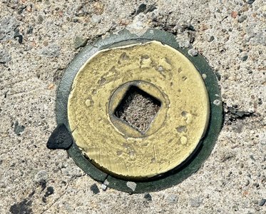 A brownish piece of sidewalks. It has dark inclusions of varying sizes. Of note is a larger black one in the top left corner. In the middle is a raised copper metal circle. In the center is a square diamond socket that goes down to the cement. Around the circle is a dark ring that slope down to the sidewalk.