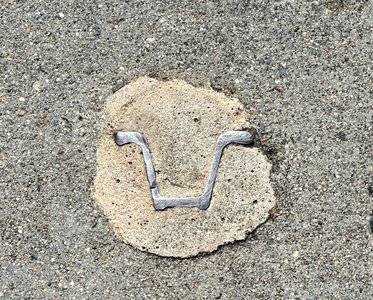 A medium gray sidewalk. In the middle is an amorphous circle of a lighter cement. In it is a thin piece of shiny metal that's been sanded down. It looks like the outline of a hat with a brim on its side that's been cut in half. It has the mood of an alien glyph of some kind.