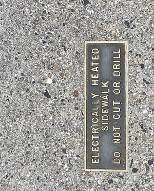 A gray sidewalk with small dark and light inclusions. A dark copper plate with a shinier raised edge and text that says, "Electrically heated sidewalk do not cut or drill"