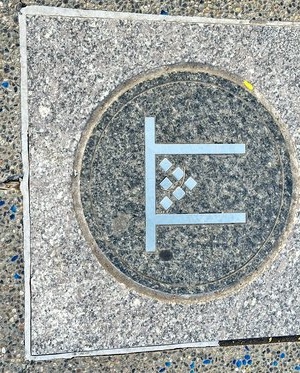 A medium gray granite looking sidewalk divided vertically into two pieces. Covering most of that is a light gray granite looking square. Inside of that is a dark granite looking circle. In the circle is the outline of the side view of a simple table--or 𝚷--with six diamonds arranged in an inverted pyramid between the table legs. The table and diamonds are a matte silver metal.